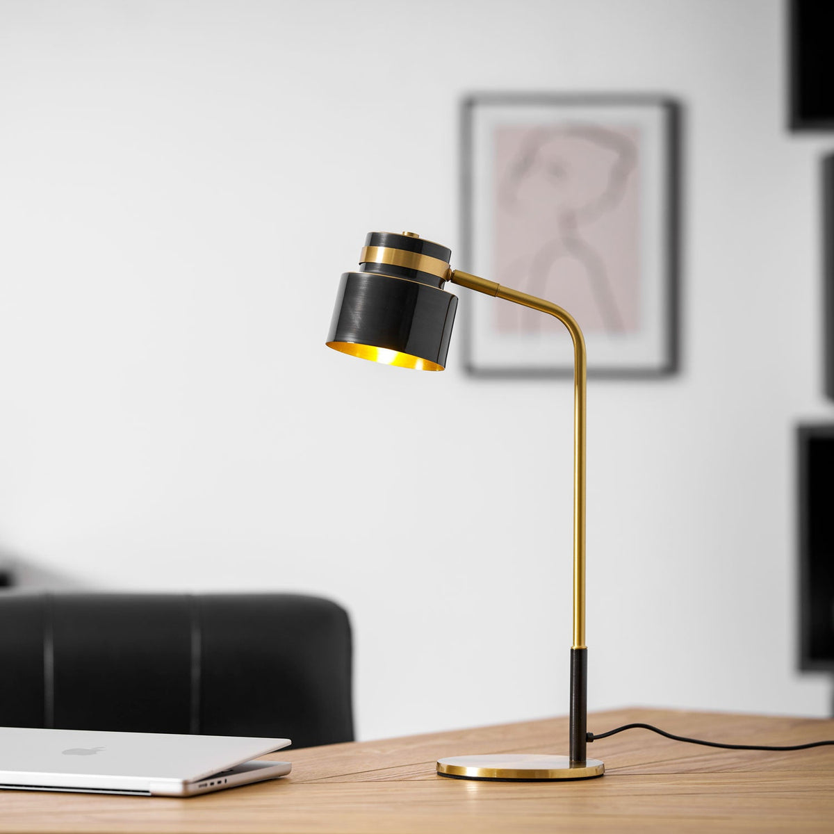 A Clérachic table lamp with its gold-lined shade tilted toward the camera, on a desk before a framed line drawing.
