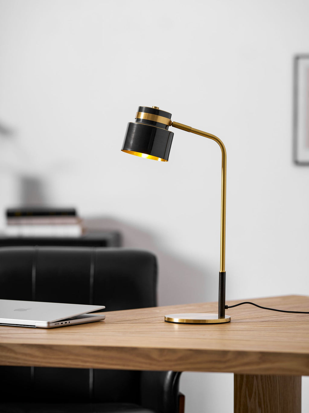 A Clérachic desk lamp on an oak table, with a laptop at the left and a black leather chair softly blurred behind.