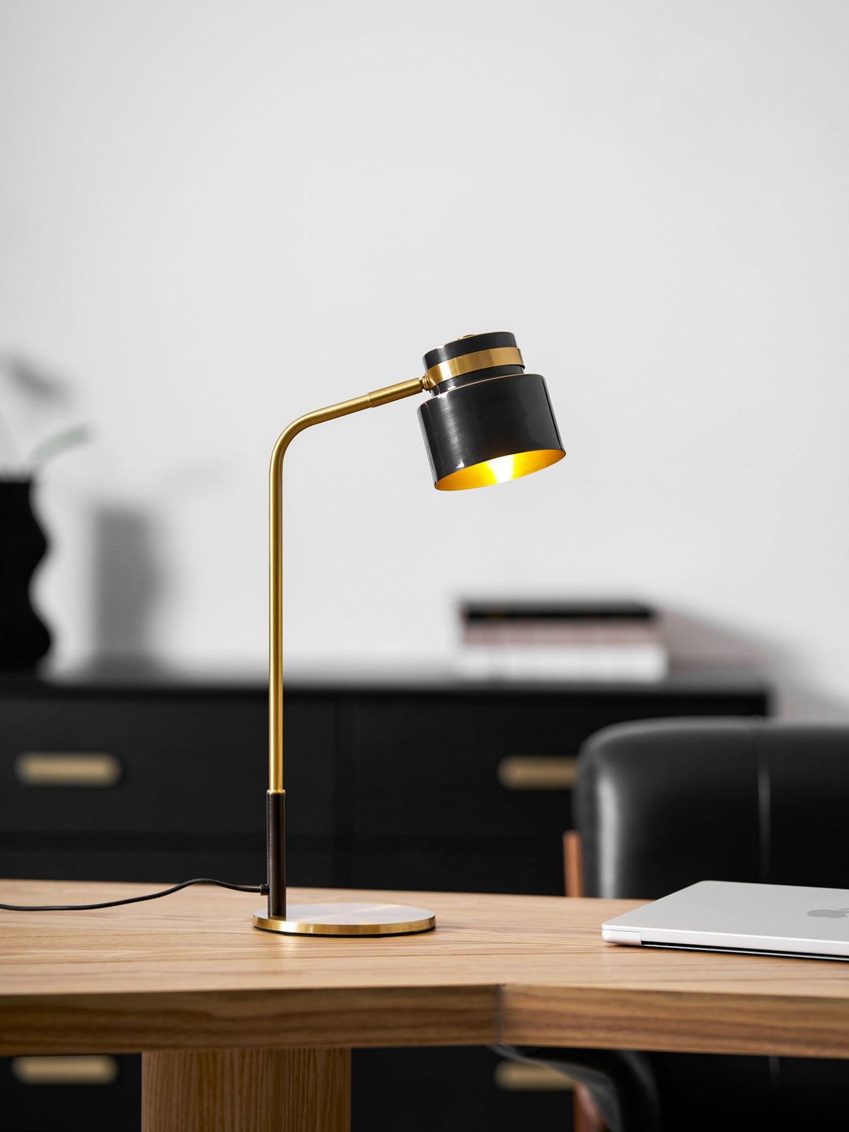 A Clérachic lamp on the oak desk with its shade angled down, in front of a black sideboard and stacked books.