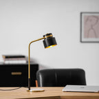 This Clérachic table lamp on a wooden desk with a black chair, a closed laptop and a framed print in a bright room.