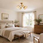 Brass Clérachic ceiling light with textured glass globes in a bedroom with a linen bed, a wood chest and bright windows.