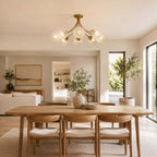Clérachic brass ceiling light over a wooden dining table with cushioned chairs, in an open room with an olive tree.