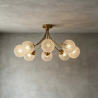 Six-globe brass Clérachic ceiling light with rippled glass, lit against a grey plaster wall and ceiling.