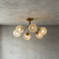 Five-globe Clérachic ceiling light with textured glass and brass arms, glowing on a grey plaster ceiling and wall.