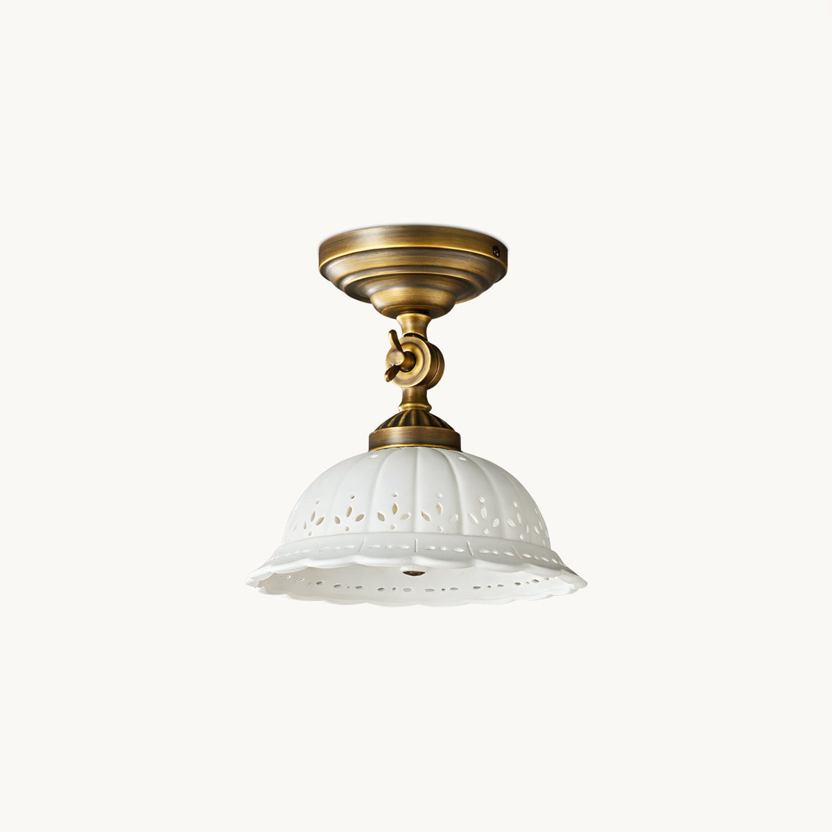 Clérachic flush-mount ceiling light with an antique brass swivel arm and a white ceramic dome shade pierced with a scalloped lace pattern, on white.