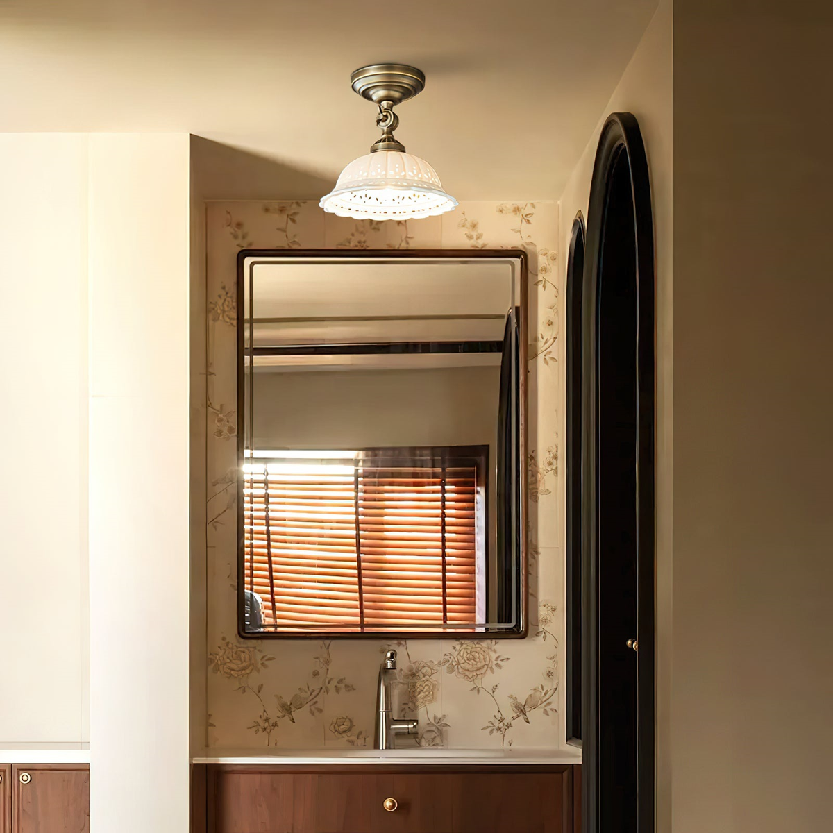 Brass and white ceramic ceiling light from Clérachic beside an arched doorway, above a washbasin with a framed mirror and floral wallpaper.