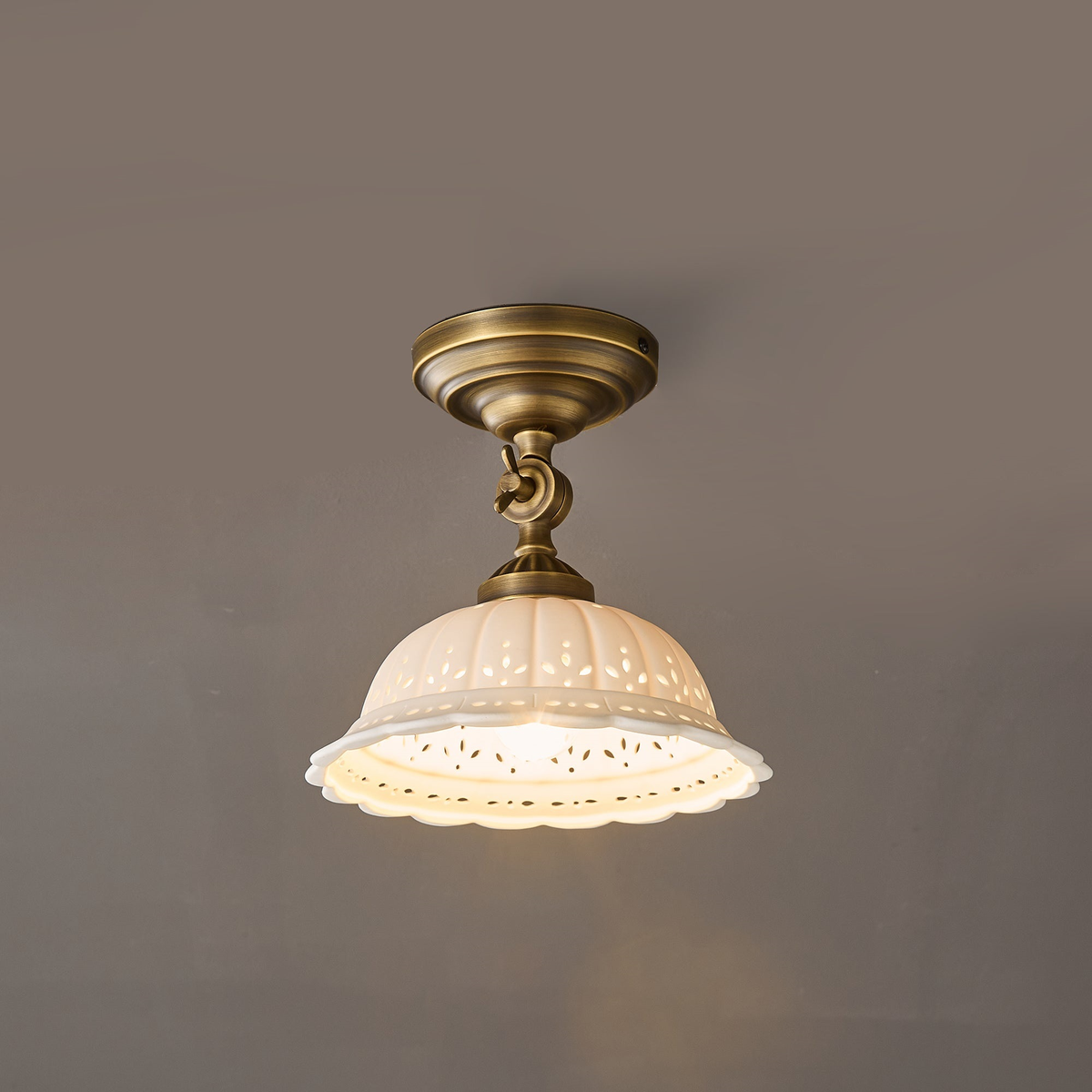 Lit Clérachic ceramic dome light on a brass swivel arm against a grey wall, the scalloped pierced rim glowing warm.