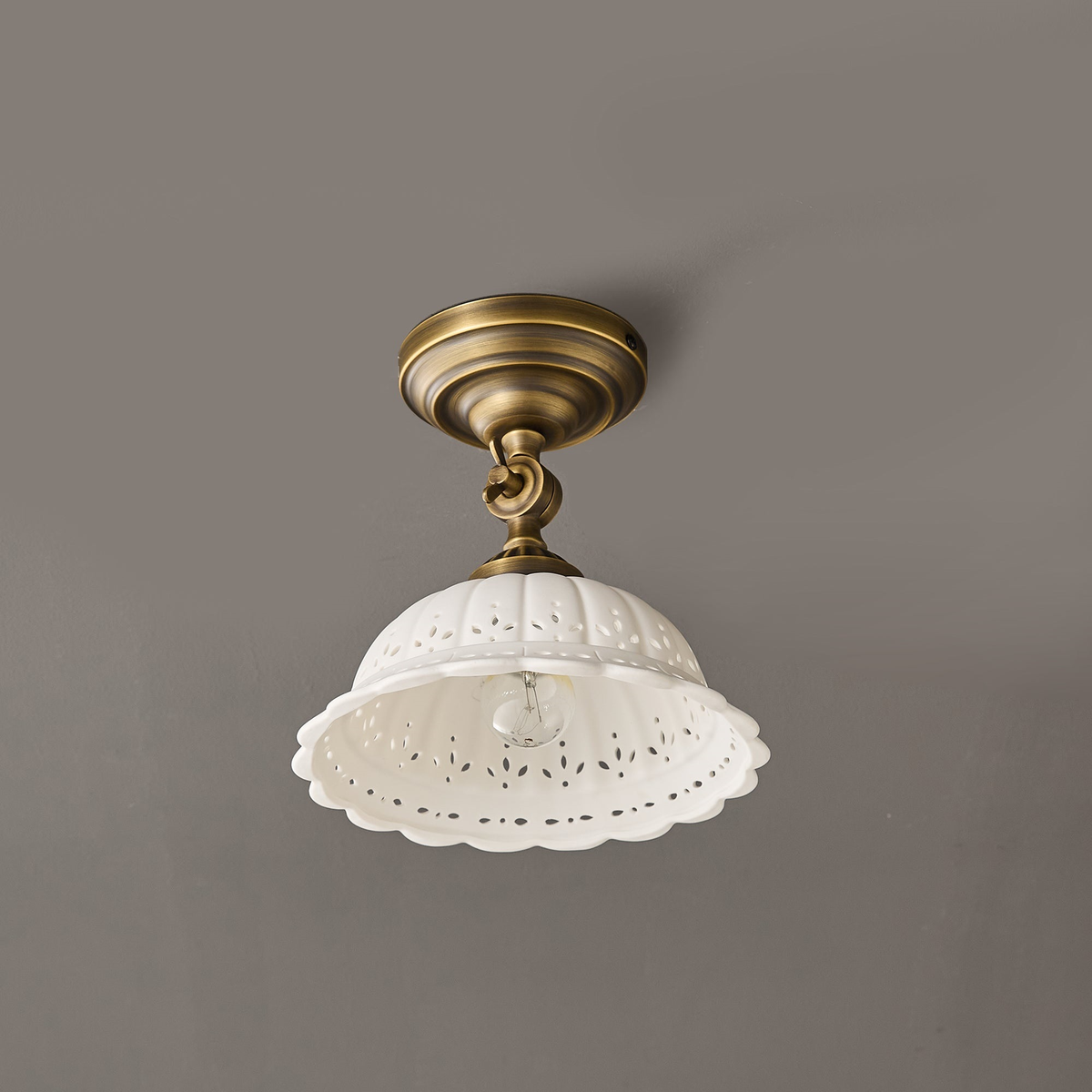 Unlit white ceramic dome on an antique brass arm, seen from below against a grey wall, with the pierced rim and bare bulb visible, from Clérachic.