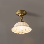 Close, lit view of a Clérachic brass ceiling light, its ceramic dome glowing warm through the cut-out pattern at the rim.