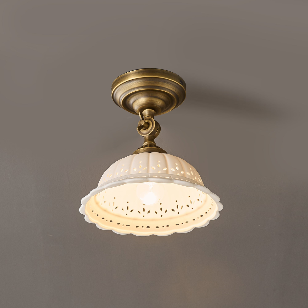 Close, lit view of a Clérachic brass ceiling light, its ceramic dome glowing warm through the cut-out pattern at the rim.