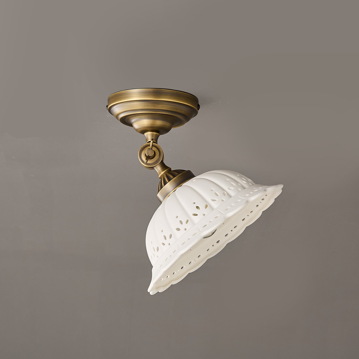 Clérachic ceiling light with its brass swivel joint angled, tilting the unlit white ceramic dome to one side against a grey wall.