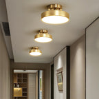 Three brass Clérachic ceiling lights stepping down a hallway, each a flat lit disc, with a wooden shelving unit at the far end.
