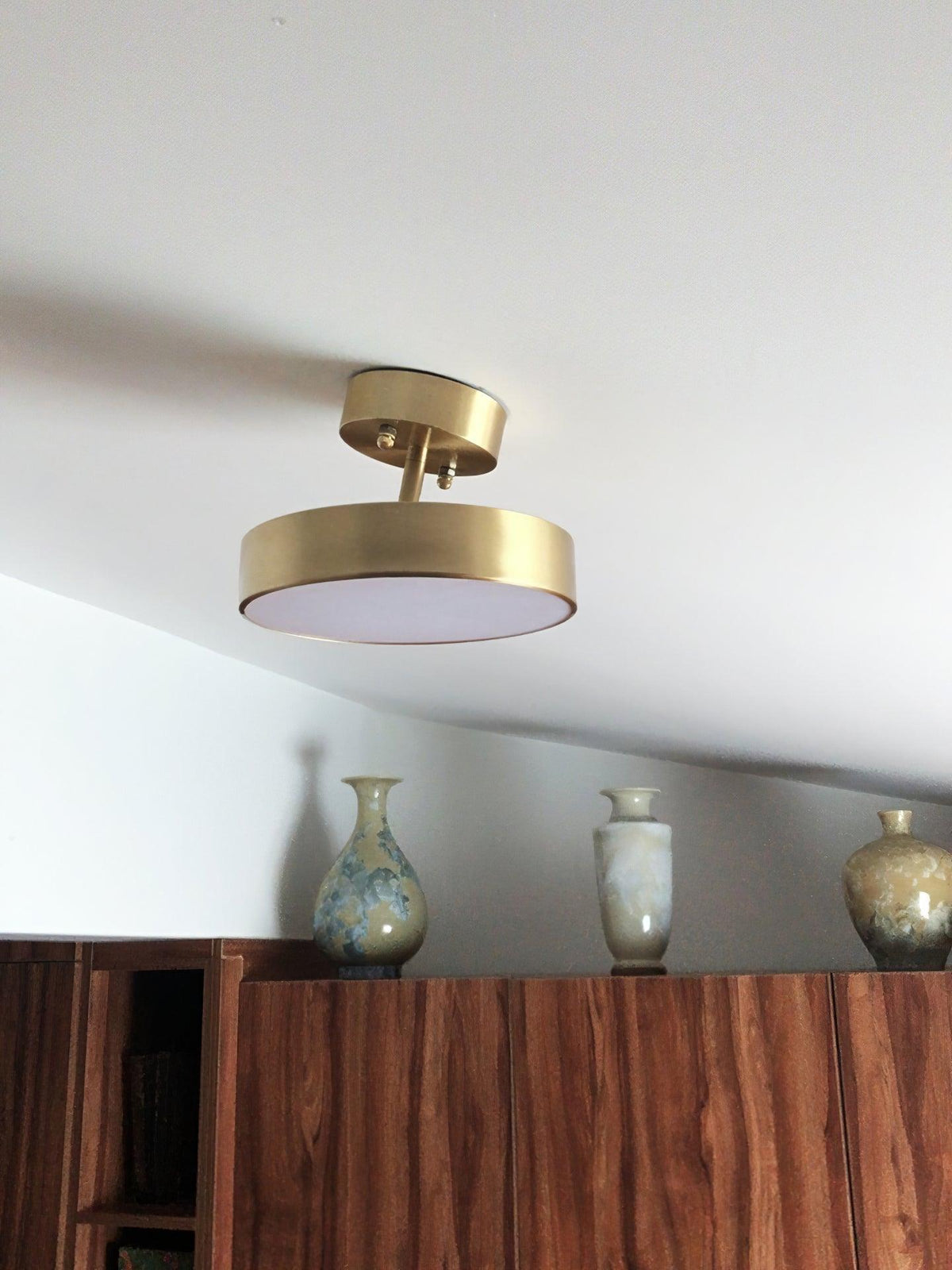 Clérachic brass ceiling light with its head tilted, set beneath a sloping ceiling above a wooden cabinet and three vases.