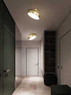 Two brass Clérachic ceiling lights, one tilted, in a hallway with a dark green door and a tall black bookshelf.