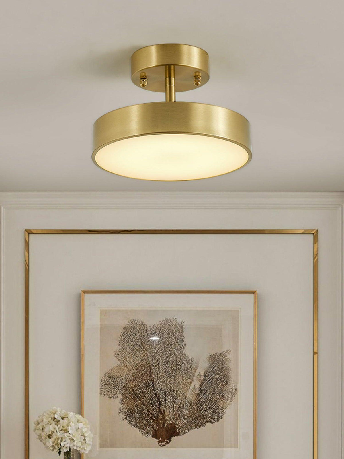 A Clérachic brass ceiling light lit above a gold-framed print of pale coral, with a bunch of white flowers at the edge.