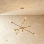 Clérachic sputnik-style chandelier in polished brass with crossed arms, hung from a plaster ceiling in studio light.