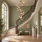 Clérachic chandelier lit in a green-walled hall with an arched window, above a dark wood staircase and a pale upholstered bench.