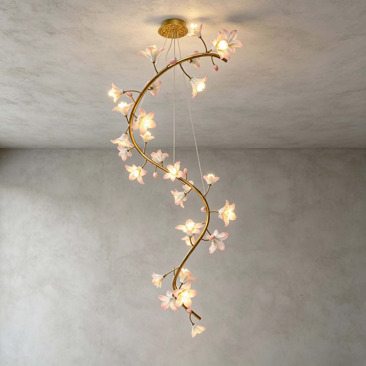 Full view of this Clérachic chandelier lit in an empty room with a grey plaster wall, the brass vine curving down from its gold canopy.