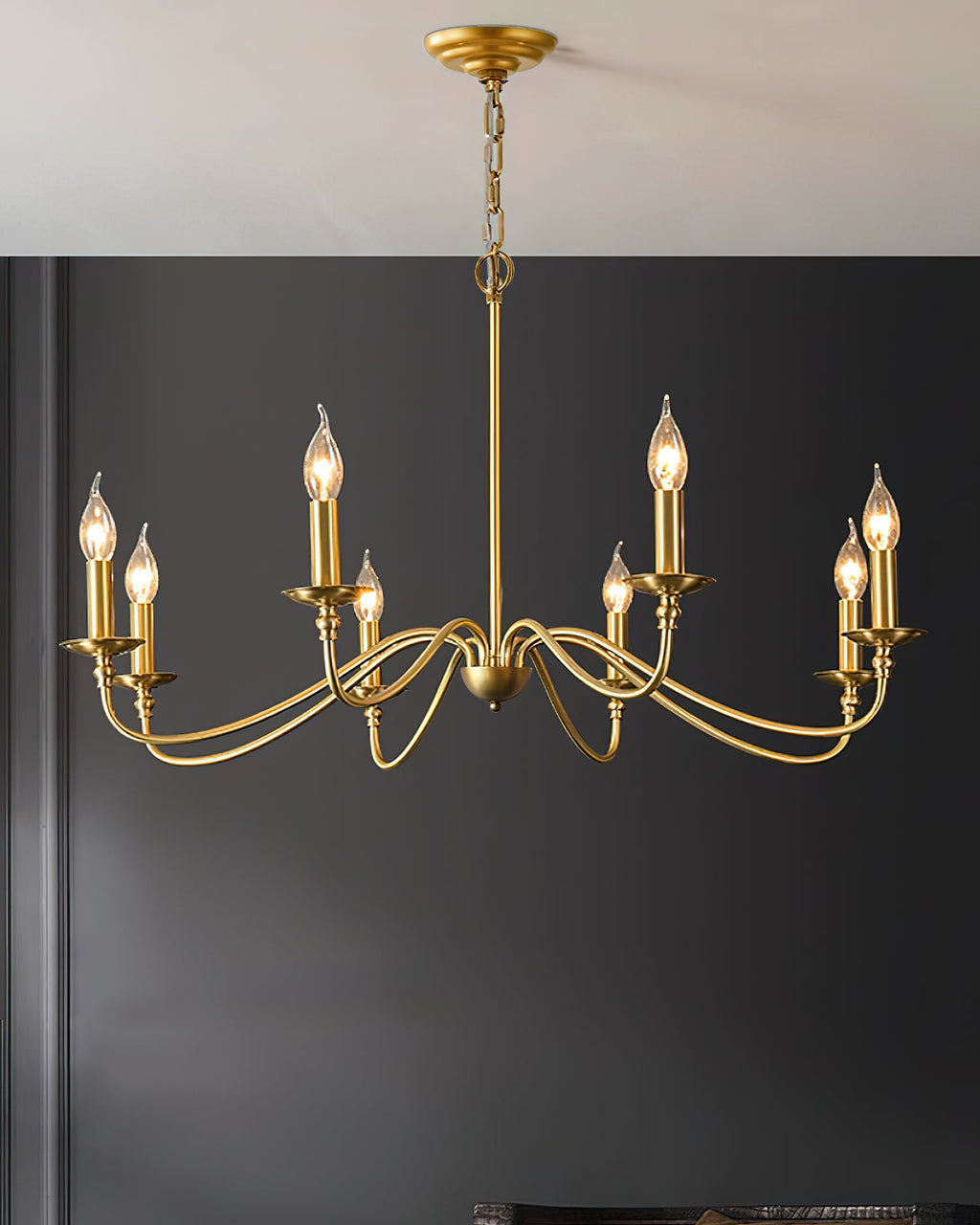 A Clérachic brass chandelier with candle bulbs hangs against a charcoal wall, from a gold ceiling plate on a chain.