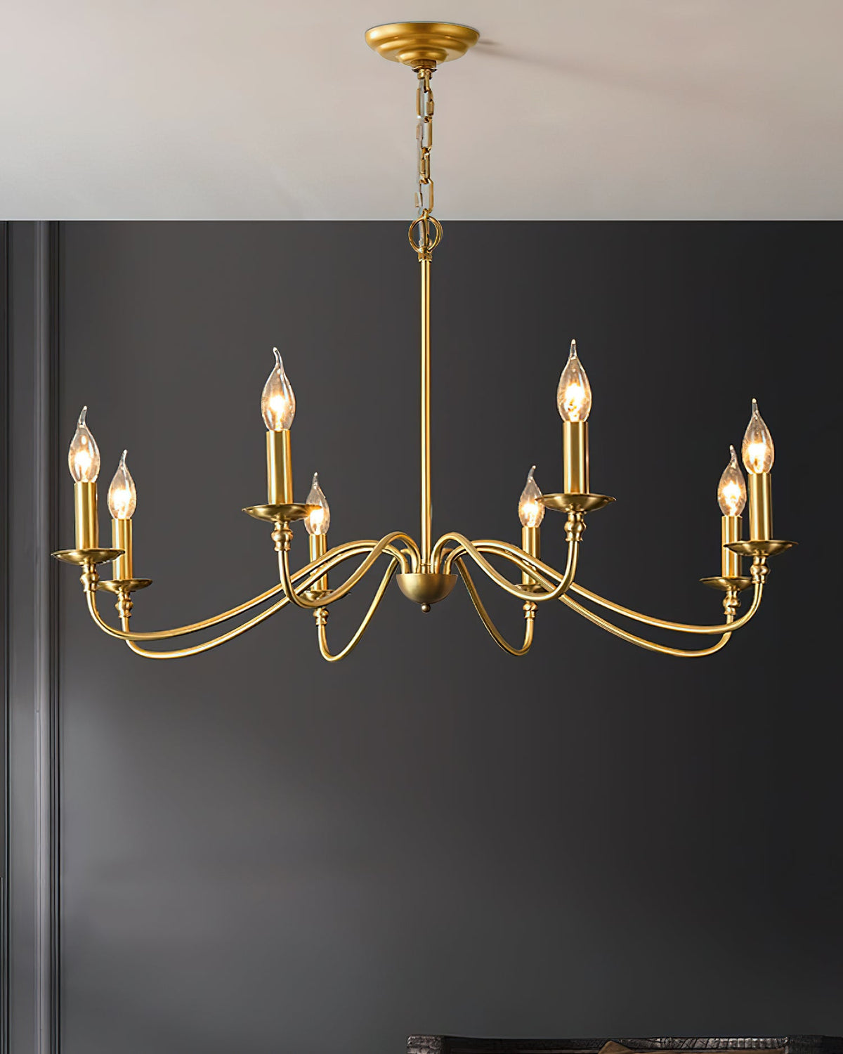 A Clérachic brass chandelier with candle bulbs hangs against a charcoal wall, from a gold ceiling plate on a chain.