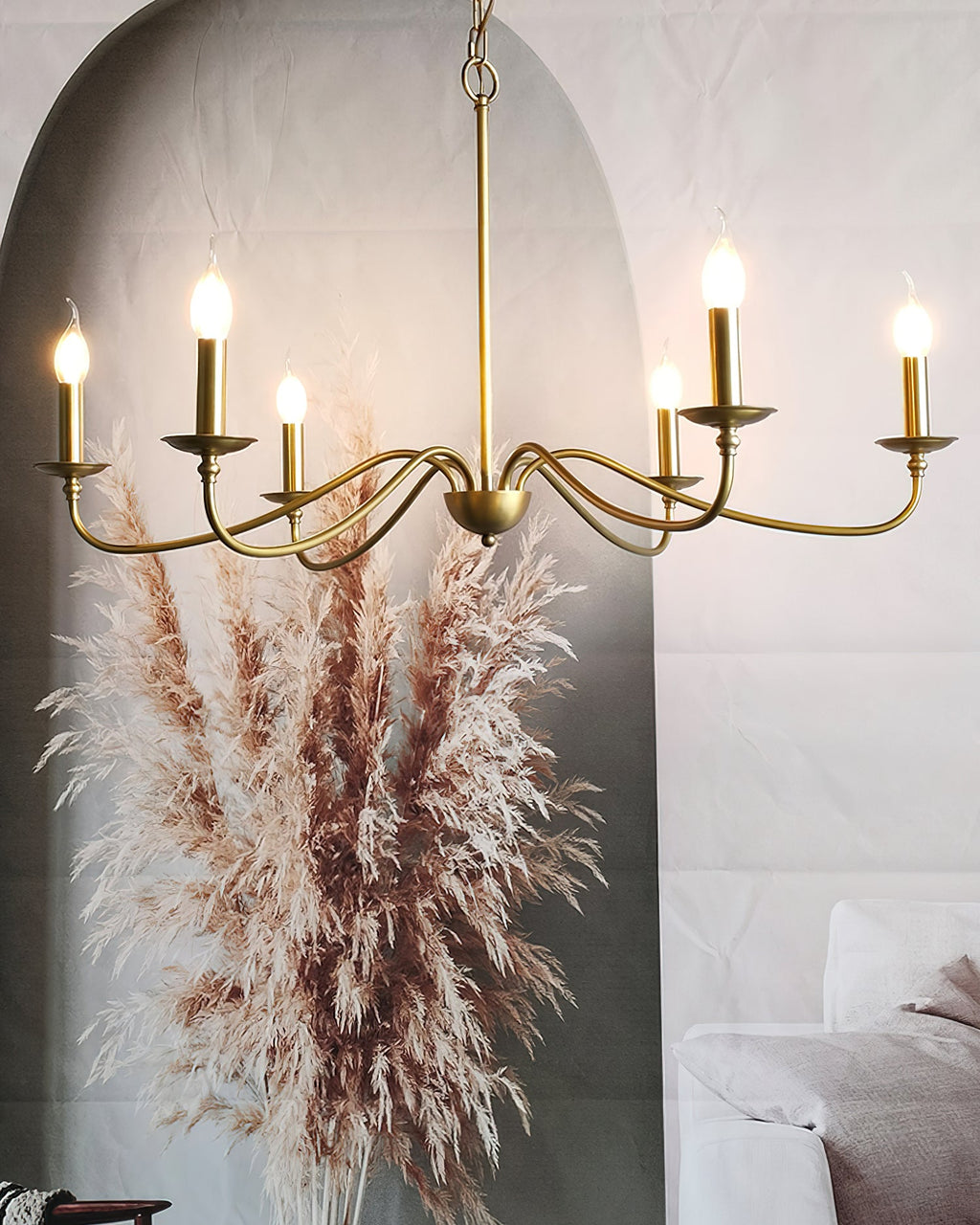 A lit Clérachic brass chandelier hangs in front of an arched wall with pampas grass and a pale sofa at the edge.