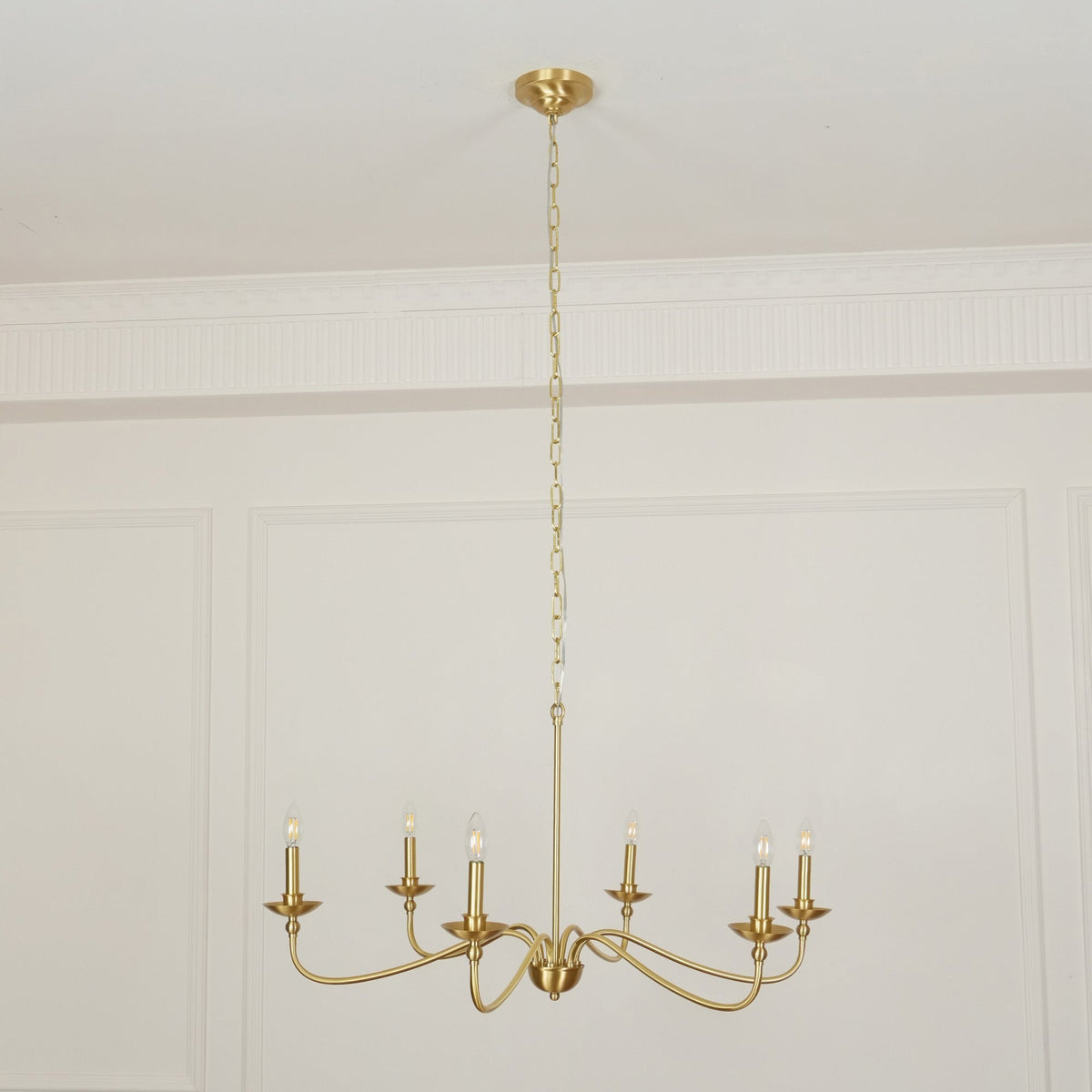 A Clérachic brass chandelier hangs low from a long chain below a panelled ceiling cornice in a pale room.