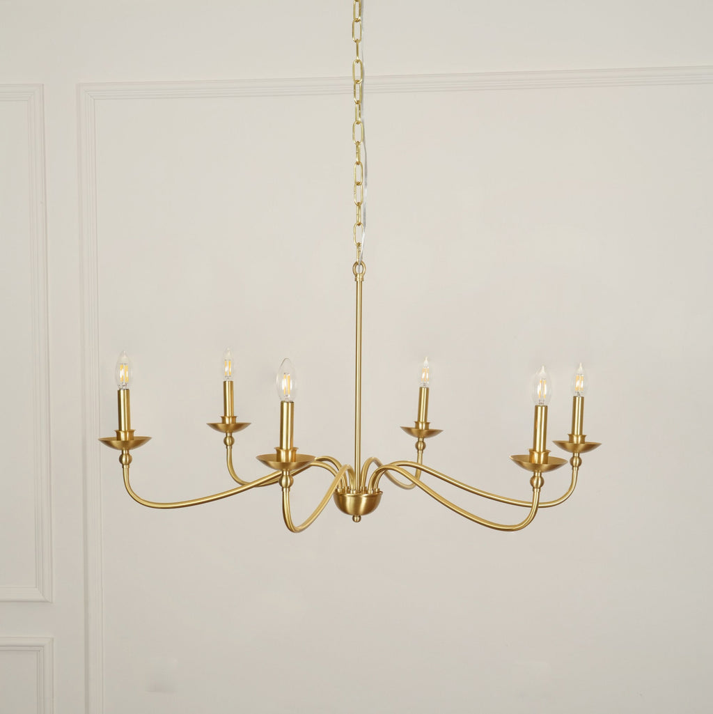 A Clérachic chandelier in a warm brass finish hangs on a chain against a white panelled wall, unlit.