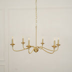 A Clérachic chandelier in a warm brass finish hangs on a chain against a white panelled wall, unlit.