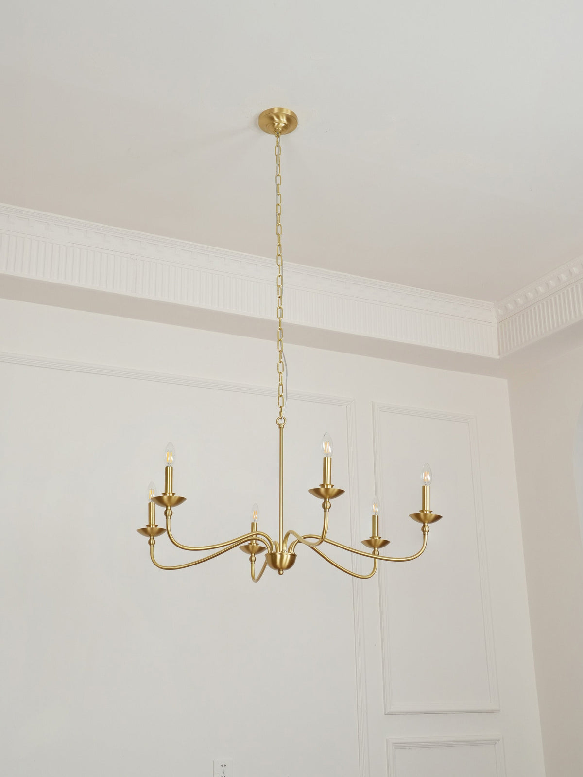 A Clérachic brass chandelier hangs from a gold ceiling plate beside a plain white wall with moulded panels.
