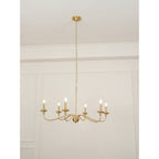 A Clérachic brass chandelier lit with candle bulbs, hung low from its chain below a cornice in a pale room.