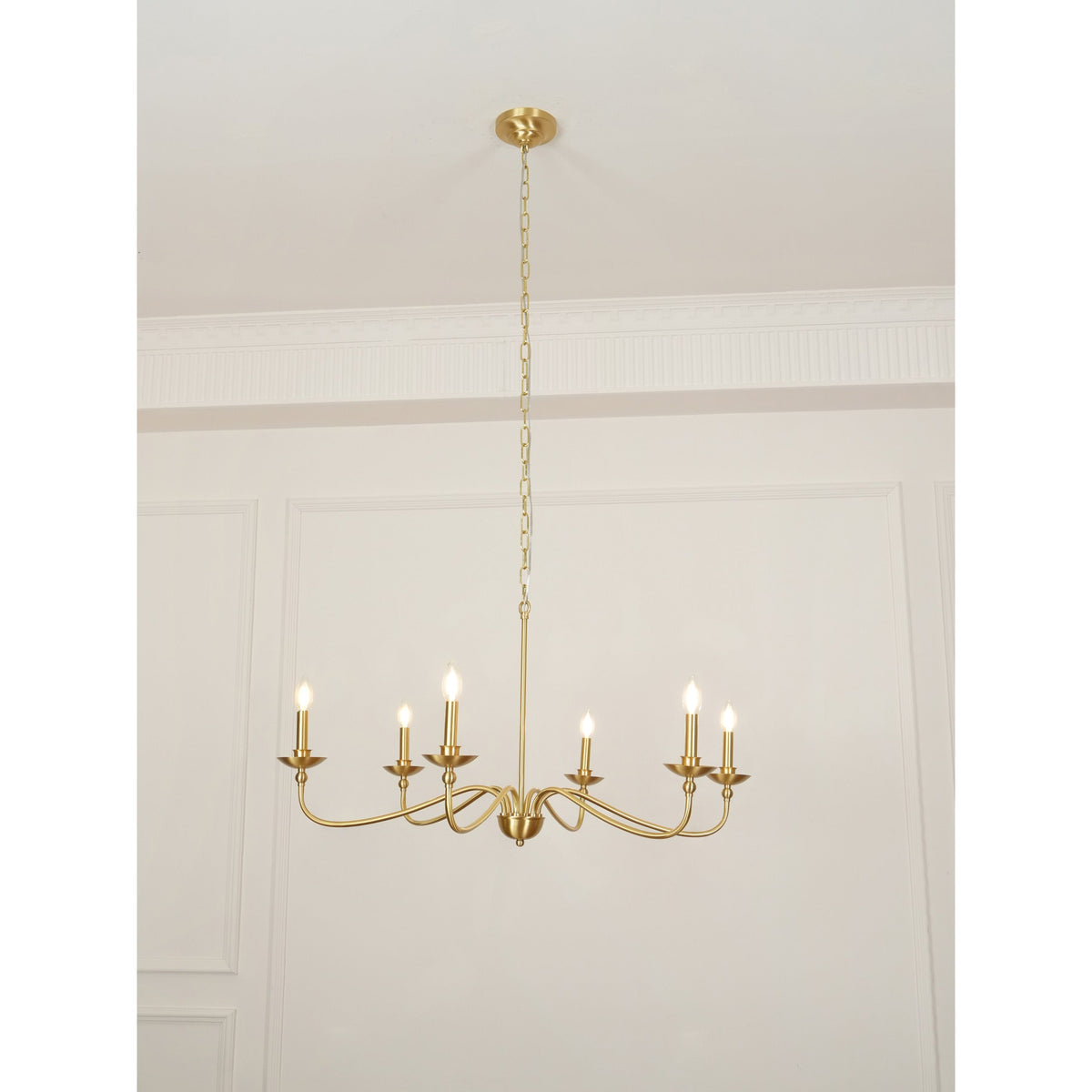 A Clérachic brass chandelier lit with candle bulbs, hung low from its chain below a cornice in a pale room.