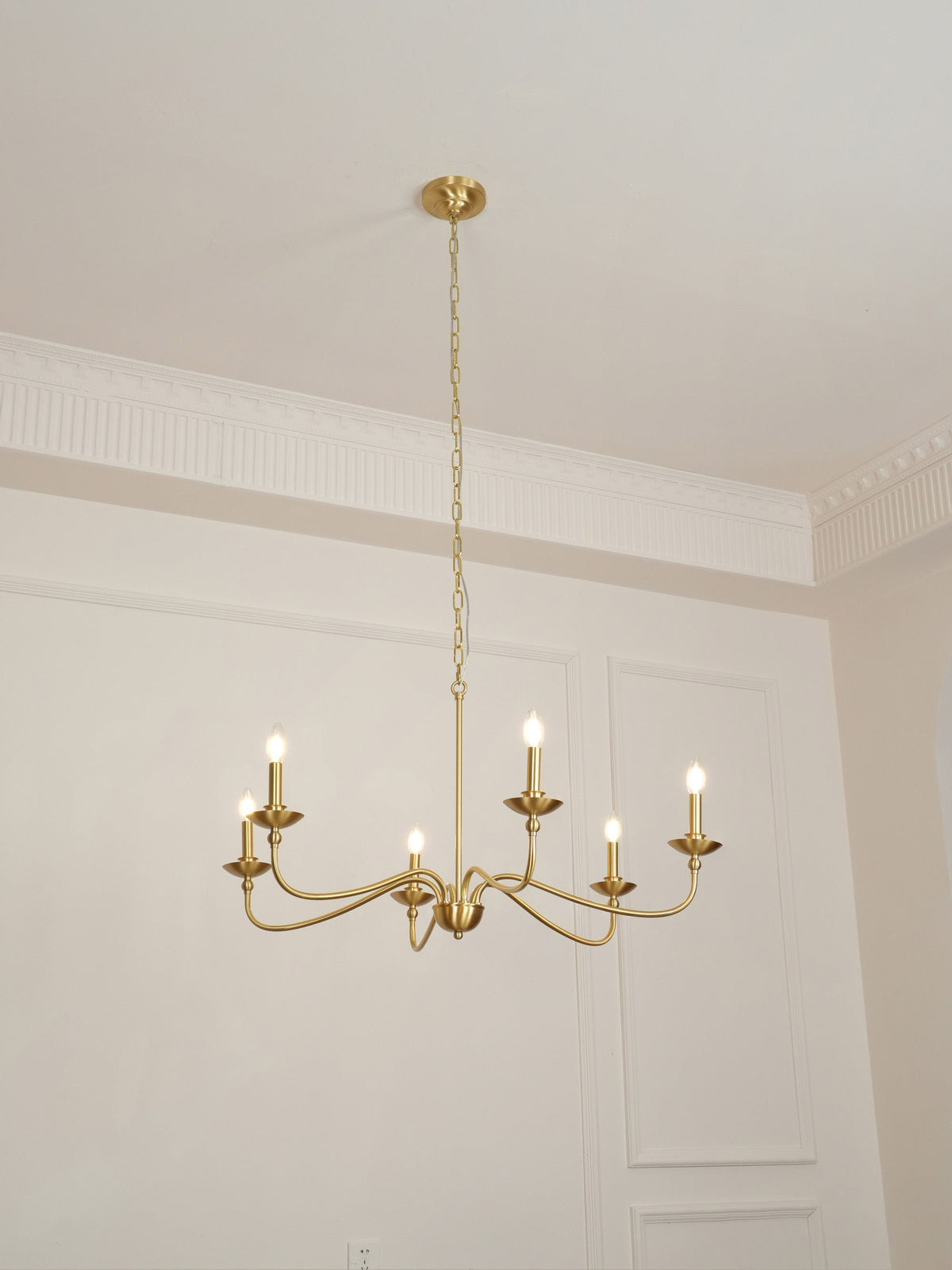 A Clérachic brass chandelier seen from below, lit, with bulbs at varied heights against pale wall panels.