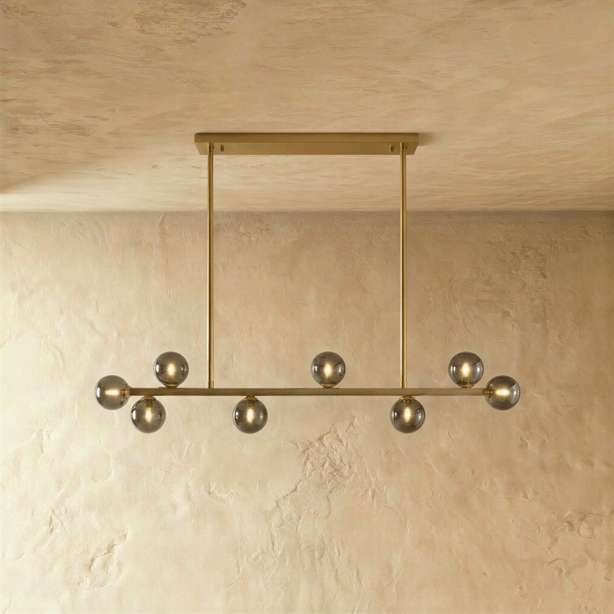 Clérachic linear chandelier hangs from a single brass horizontal bar plate, smokey grey glass globes unlit.