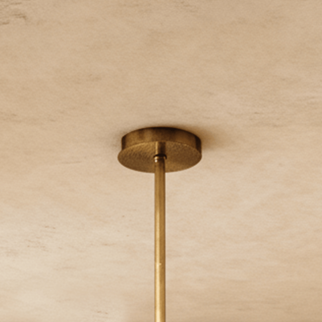 Clérachic chandelier's round brass ceiling canopy and slim brass rod seen close up against a soft limewash ceiling.