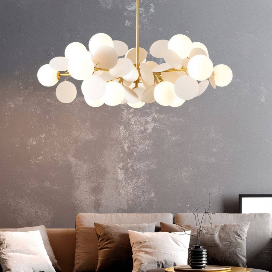 Clérachic wide chandelier of frosted glass globes and cream discs on brass arms, above a sofa and grey wall.