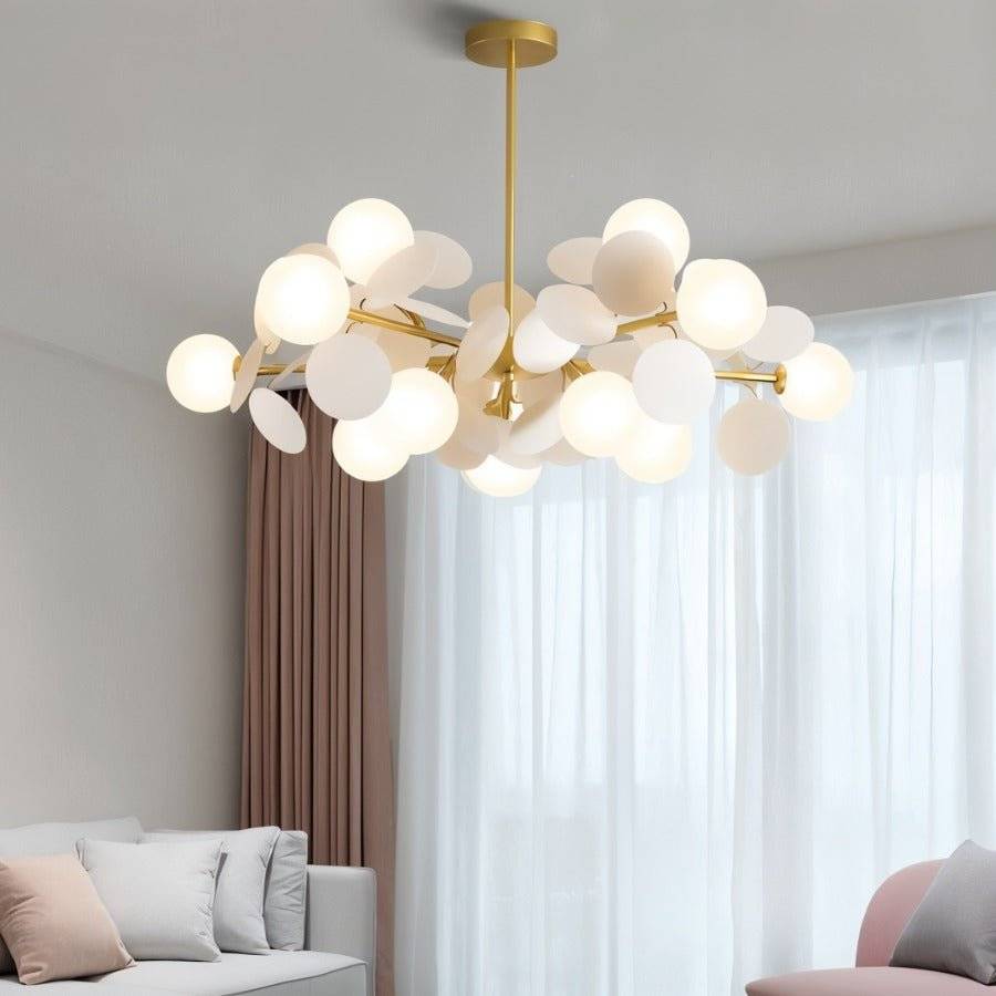 Clérachic wide chandelier of frosted globes and white discs on brass arms, in front of sheer curtains.