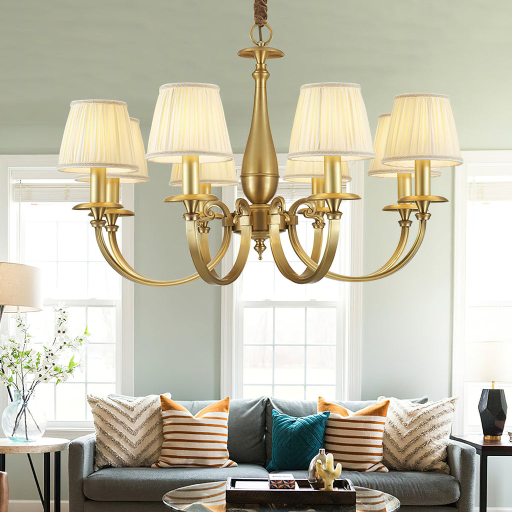 A Clérachic brass chandelier with pleated white shades hangs in a living room with sage walls, above a grey sofa and a glass coffee table.