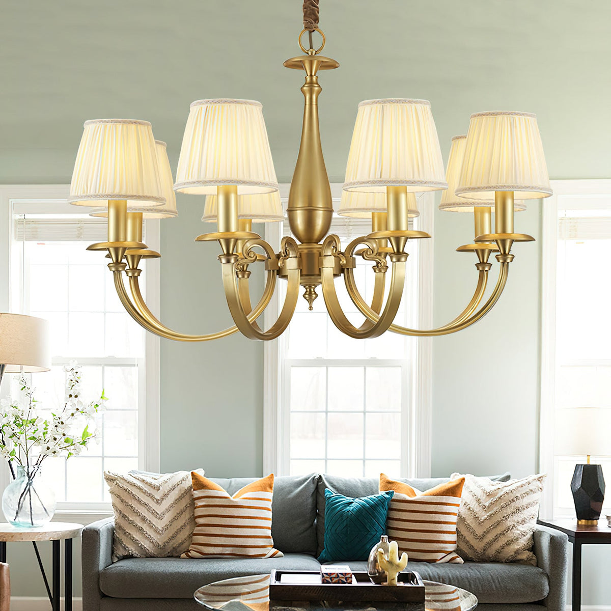 A Clérachic brass chandelier with pleated white shades hangs in a living room with sage walls, above a grey sofa and a glass coffee table.