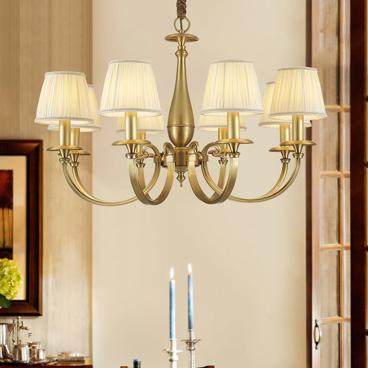 Lit Clérachic brass chandelier in a dining room, above a table set with tall blue candles, a framed picture to the left.