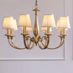 This Clérachic brass chandelier with pleated fabric shades hangs against a lilac-grey panelled wall, its antique brass finish glowing.