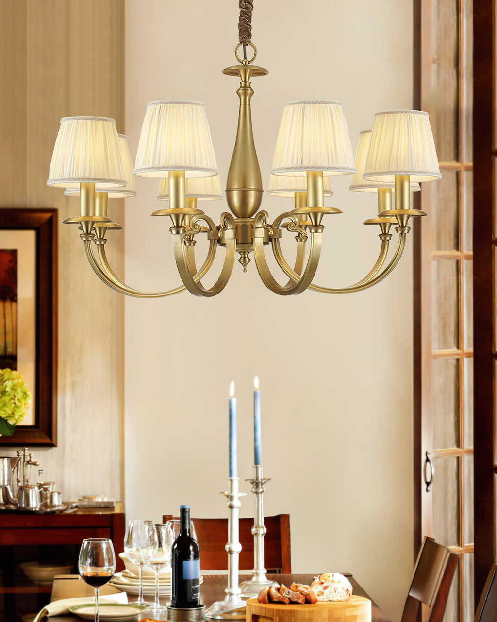 A Clérachic brass chandelier above a laid dining table, with wine glasses, a bottle, candlesticks and a bread board below it.