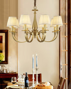 A Clérachic brass chandelier above a laid dining table, with wine glasses, a bottle, candlesticks and a bread board below it.