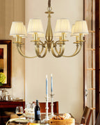 A Clérachic brass chandelier lit in a dining room, with a set table, candlesticks and a dark wood door frame behind it.