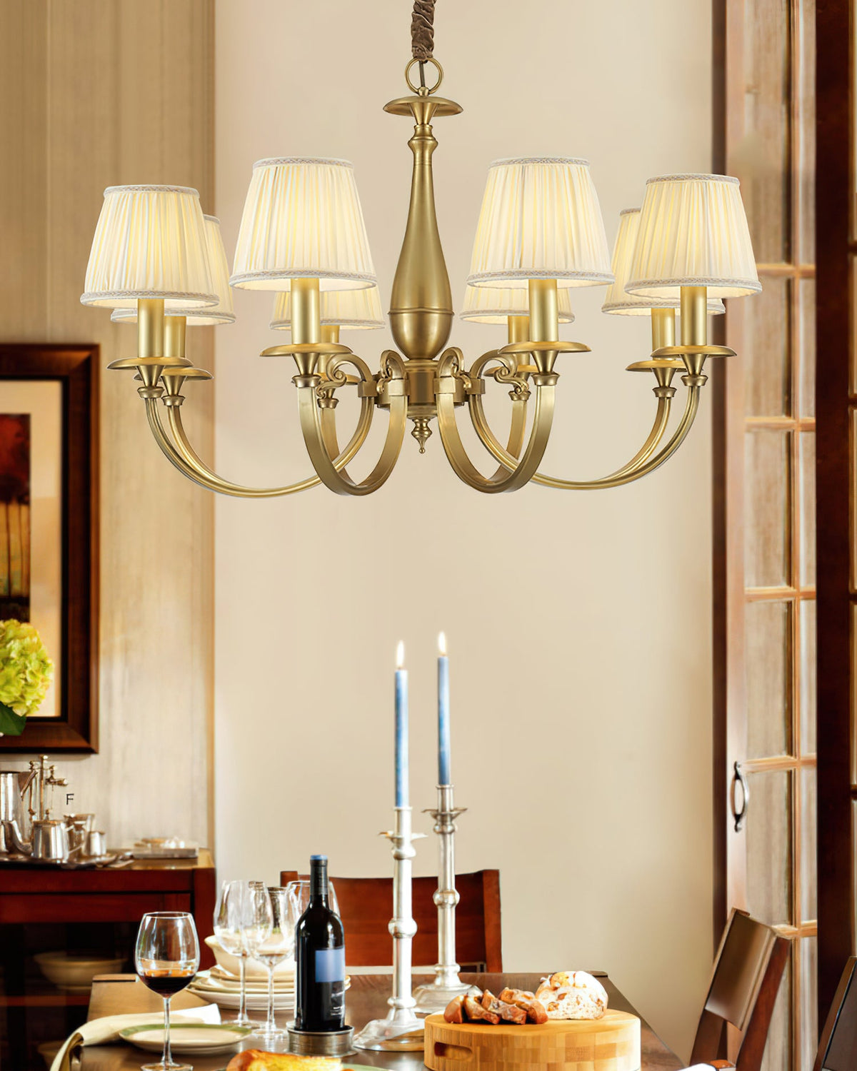 A Clérachic brass chandelier lit in a dining room, with a set table, candlesticks and a dark wood door frame behind it.