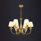 A Clérachic brass chandelier with a six-arm spread on a dark backdrop, pleated white shades lit, hung from a chain.