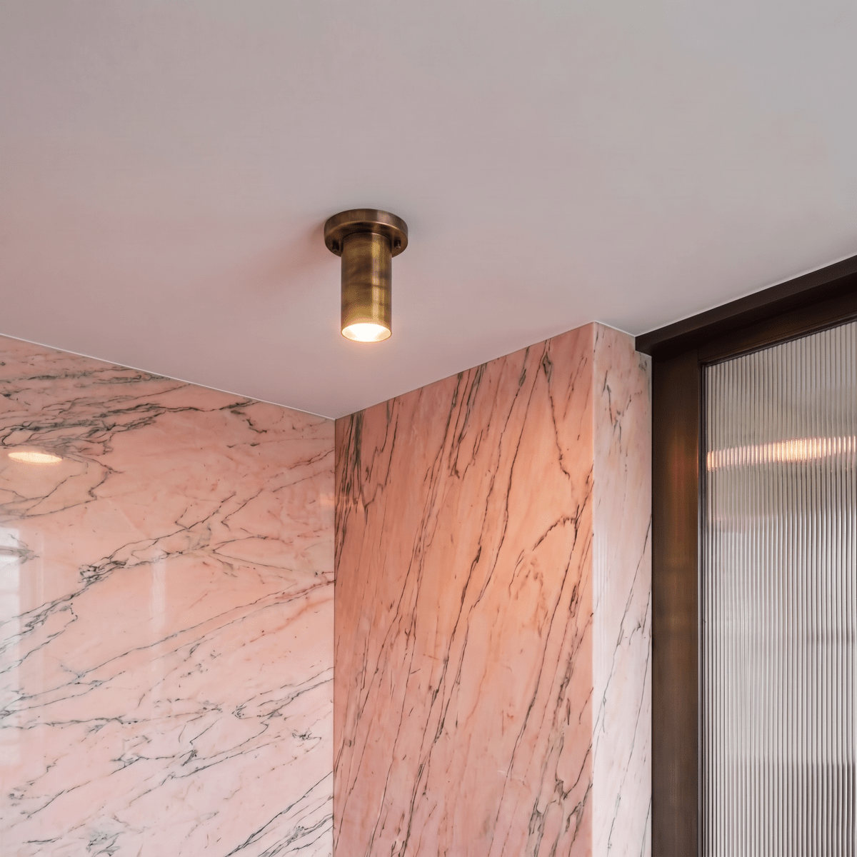 A vintage brass Clérachic ceiling light lights a pink marble corner beside a fluted glass door.