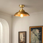A Clérachic brass dome ceiling light close to a cornice in a cream room, with an arched window and gilt-framed pictures below.