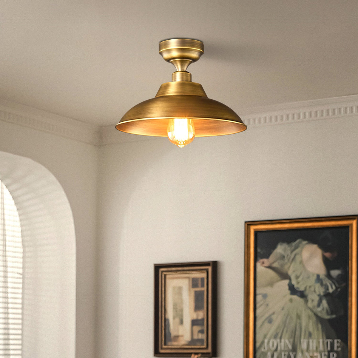 A Clérachic brass dome ceiling light close to a cornice in a cream room, with an arched window and gilt-framed pictures below.