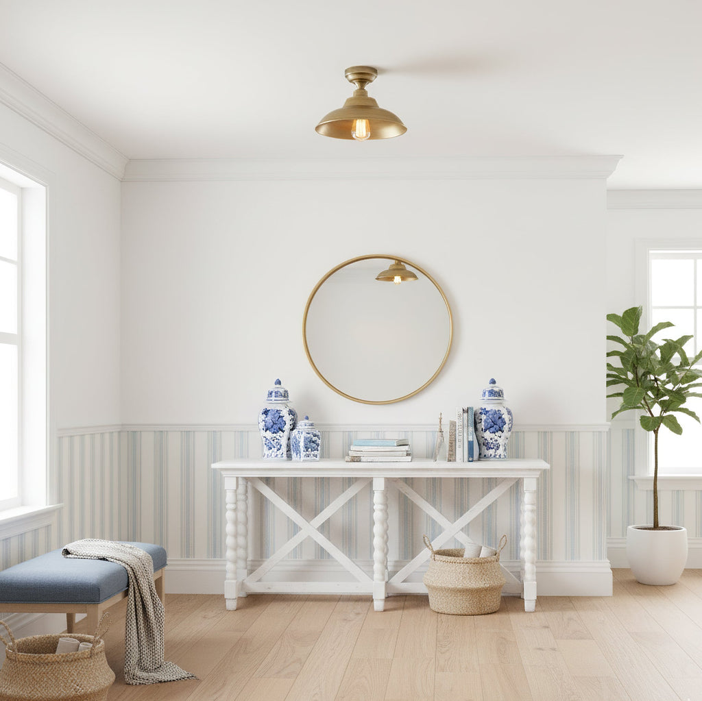 A Clérachic brass dome ceiling light in a bright white room, above a round gold mirror, a white console table and blue-and-white vases.
