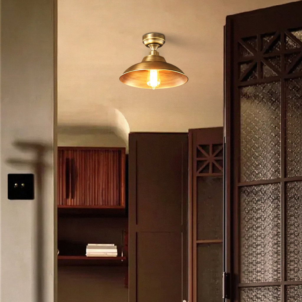 Close view of a lit Clérachic brass dome ceiling light, its warm glow inside the shade, beside dark carved wooden doors.
